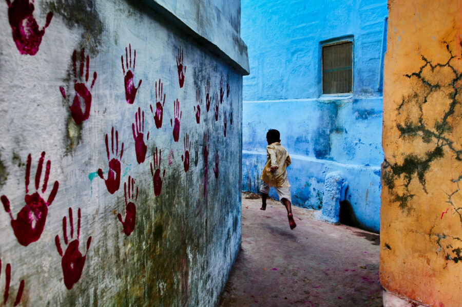 back of child running through alley. Wall on left side covered with red handprints.