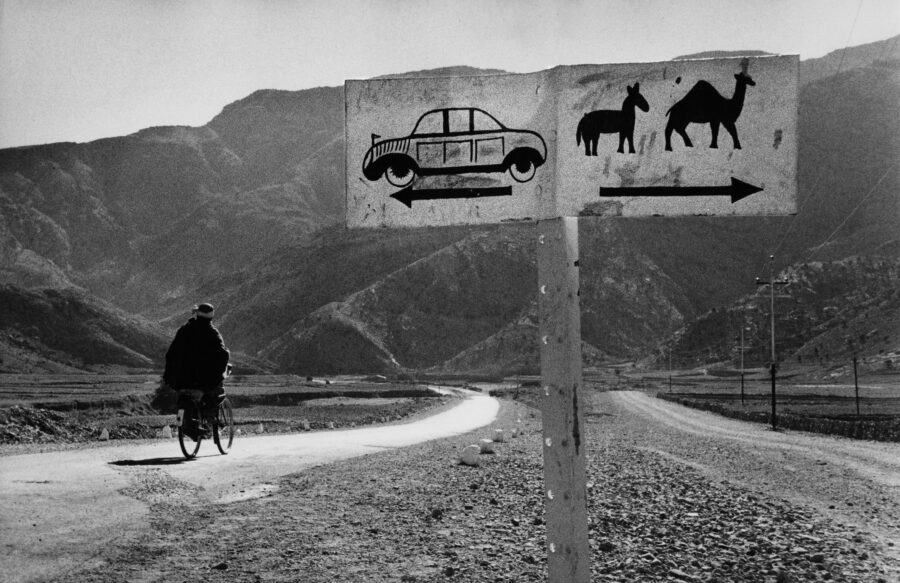 Black-and-white photograph of figure riding bicycle with mountains in background. Sign to figure's left has a drawing of a car and an arrow pointing left and a drawing of a horse and a camel with an arrow pointing right.