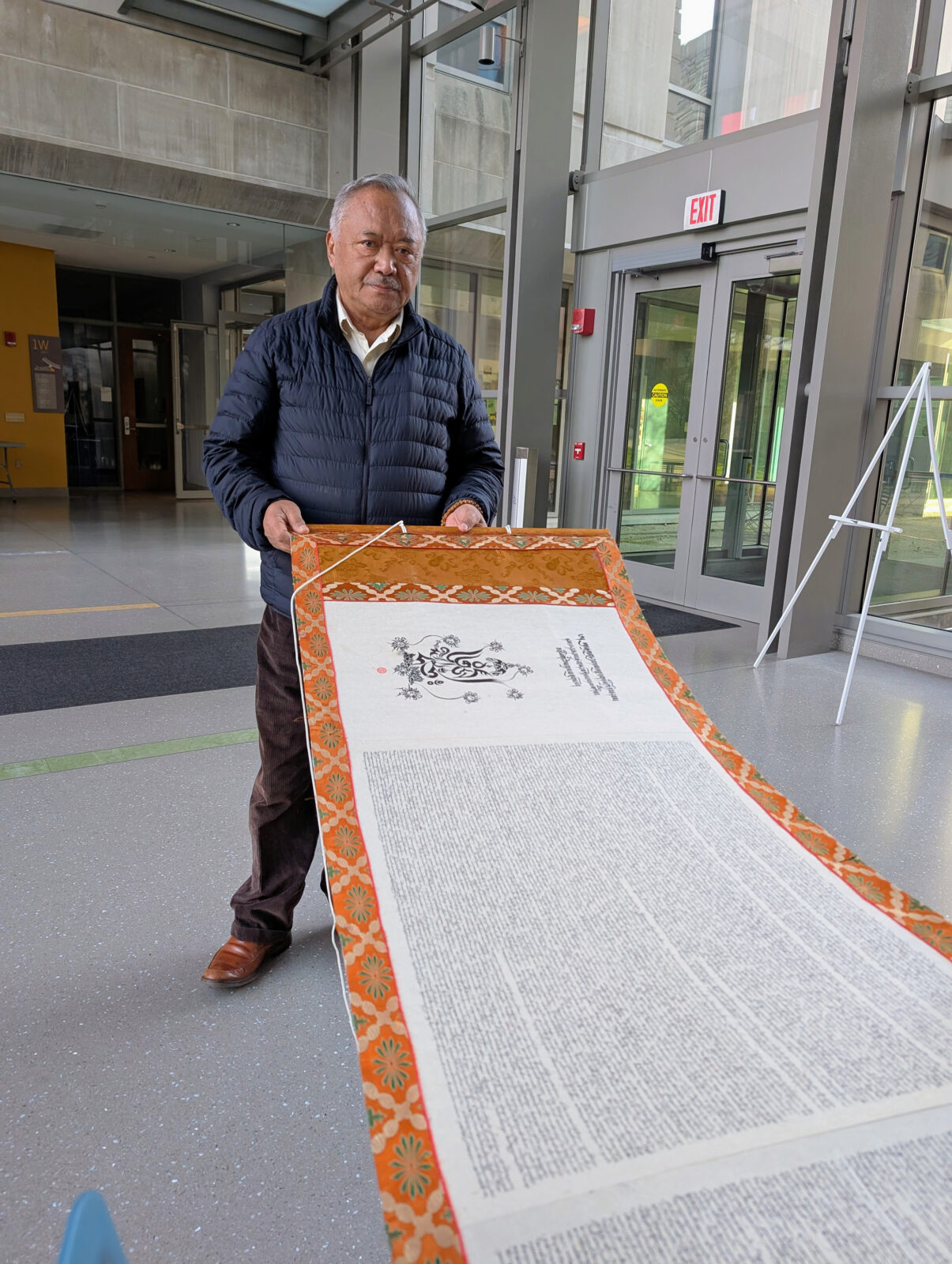 An adult man holds up one end of a large scroll of Pema Khatang with an orange border