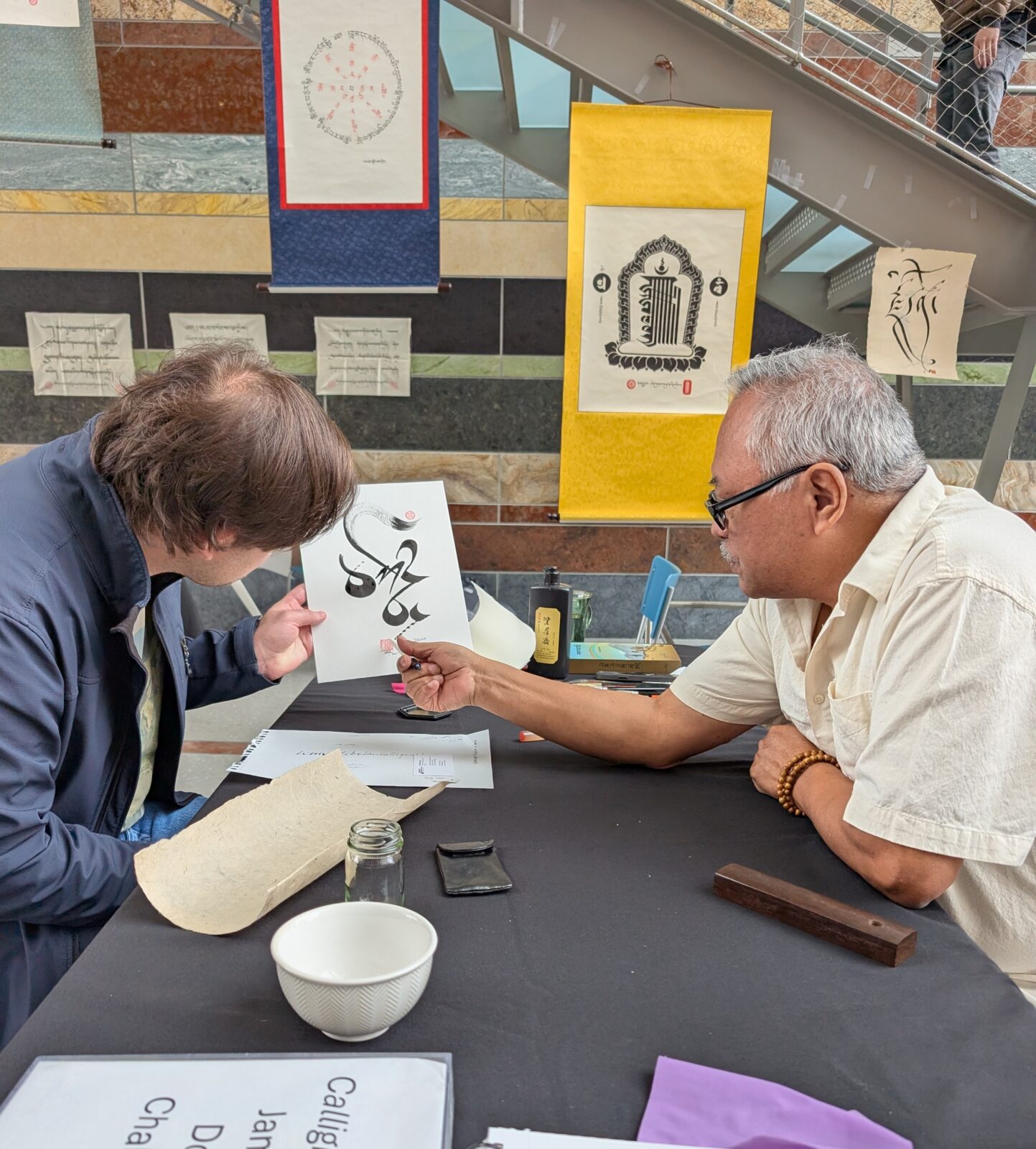 An adult man with grey hair and black glasses hands a paper with Tibetan calligraphy to another adult
