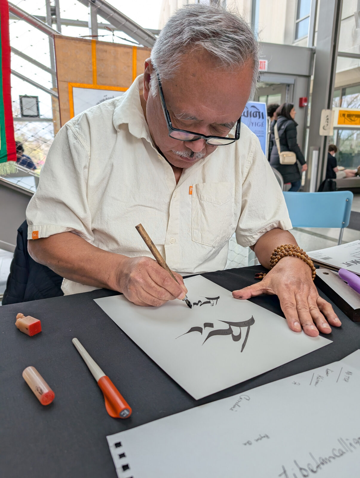 An adult man with grey hair and black glasses sits at a table writing calligraphy with a brush