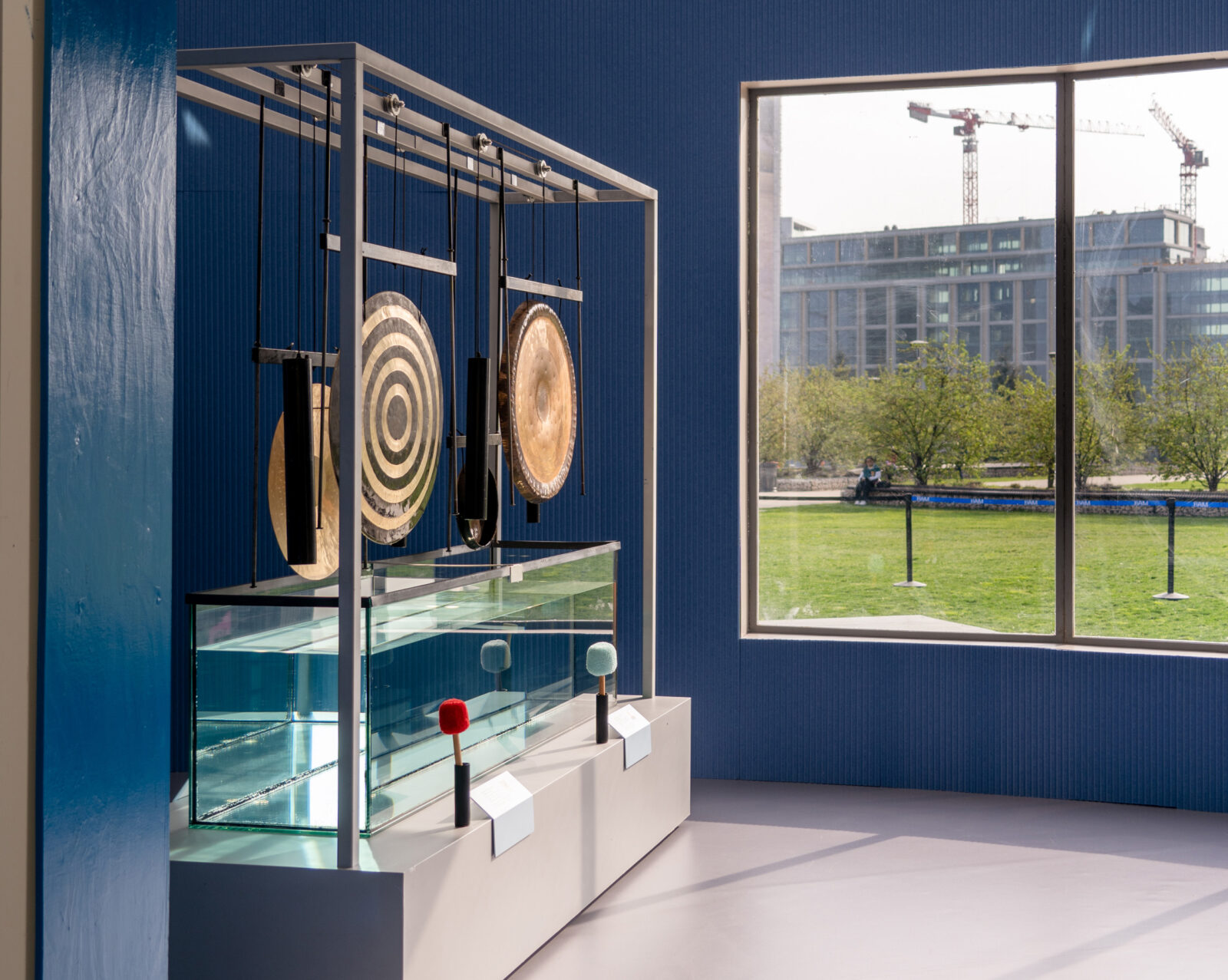 Three large gongs suspended above tank of water in dark blue blue with large window looking out on grassy field.