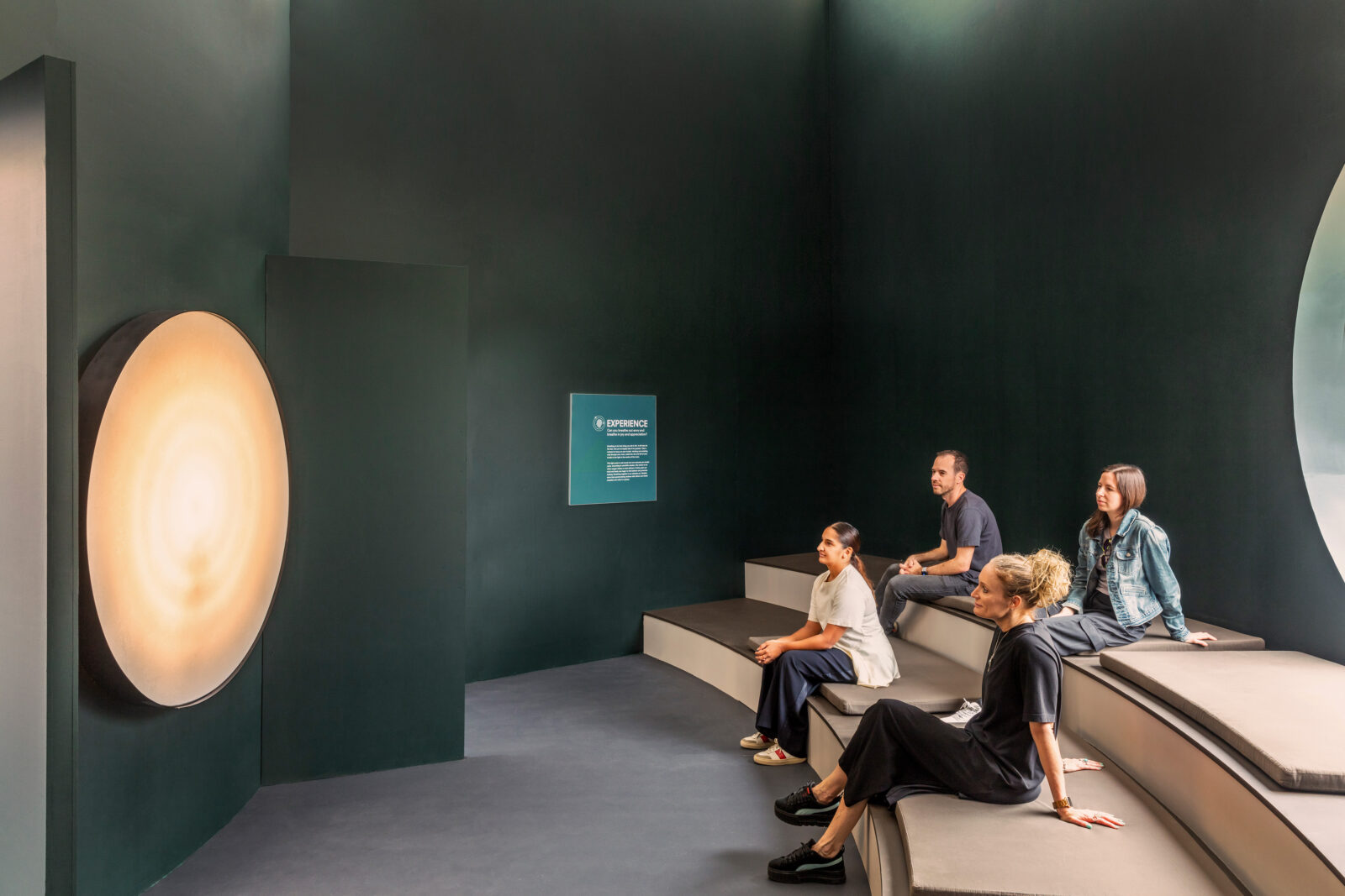 Four adults seated on wide steps in room with dark walls, looking at glowing circular object on wall.