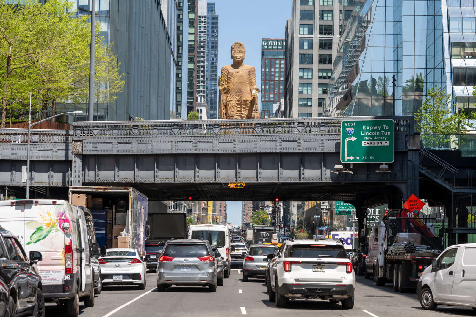 A Bamiyan Buddha sculpture with gold hands on a bridge above a street in New York City