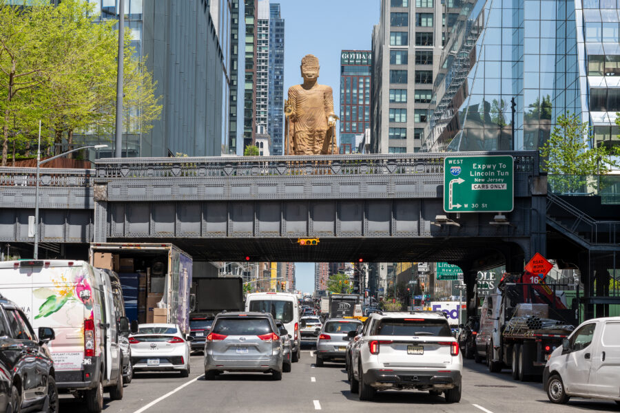 A Bamiyan Buddha sculpture with gold hands on a bridge above a street in New York City