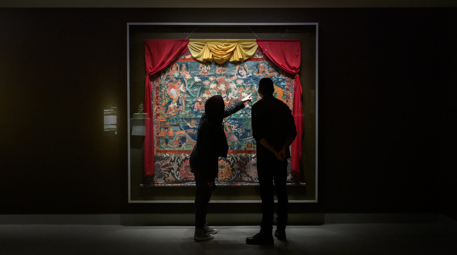 Visitors in museum space looking at large thangka painting, pointing.