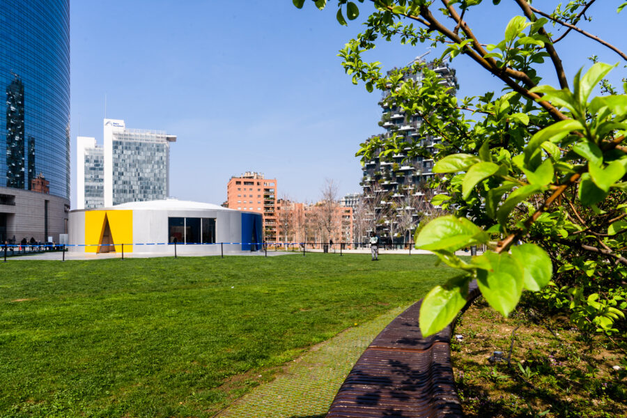 Short, gray circular building with triangular entrances situated on a grassy field. Tree branches in the foreground and skyscrapers in the background.