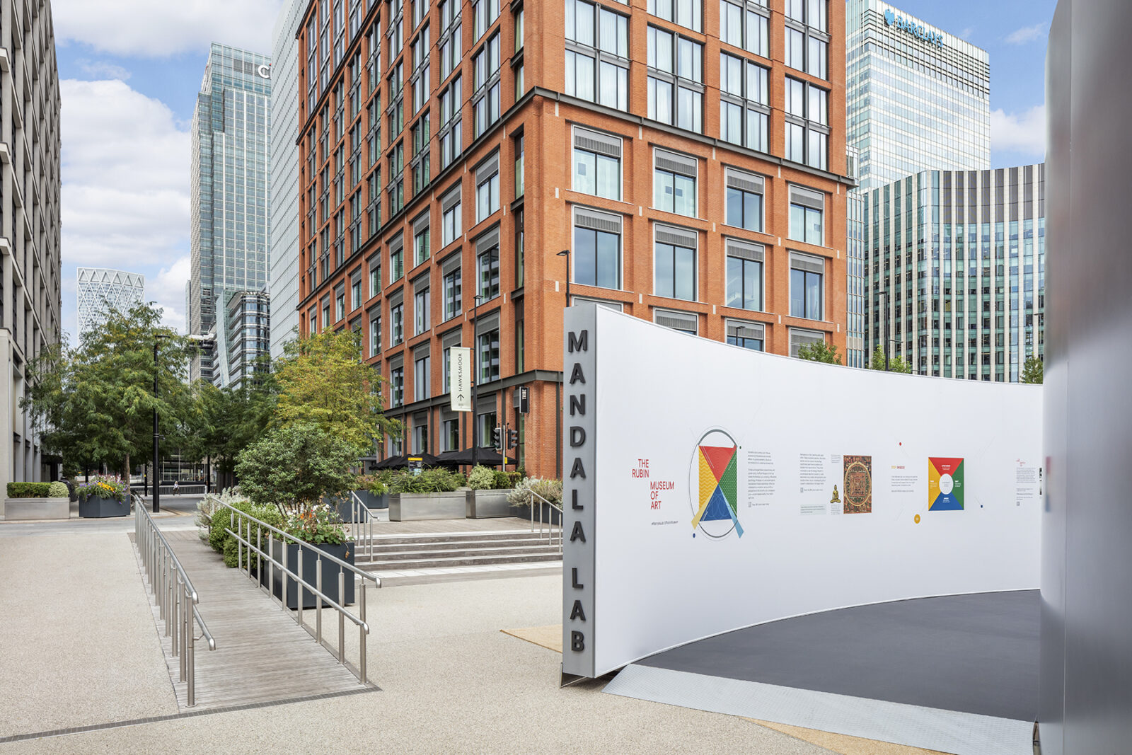 Curved entrance to structure with horizontal black letters reading "Mandala Lab" on edge of wall. Skyscrapers in the background.