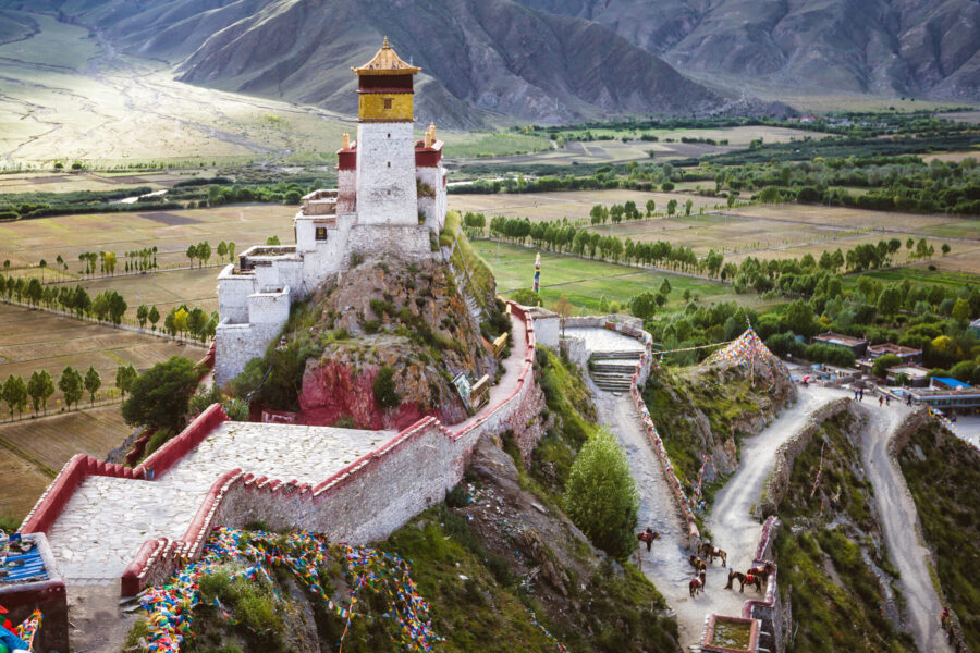 Large white brick building with gold roof perched on hill surrounded by white and red walls. Fields, trees, and mountains in background