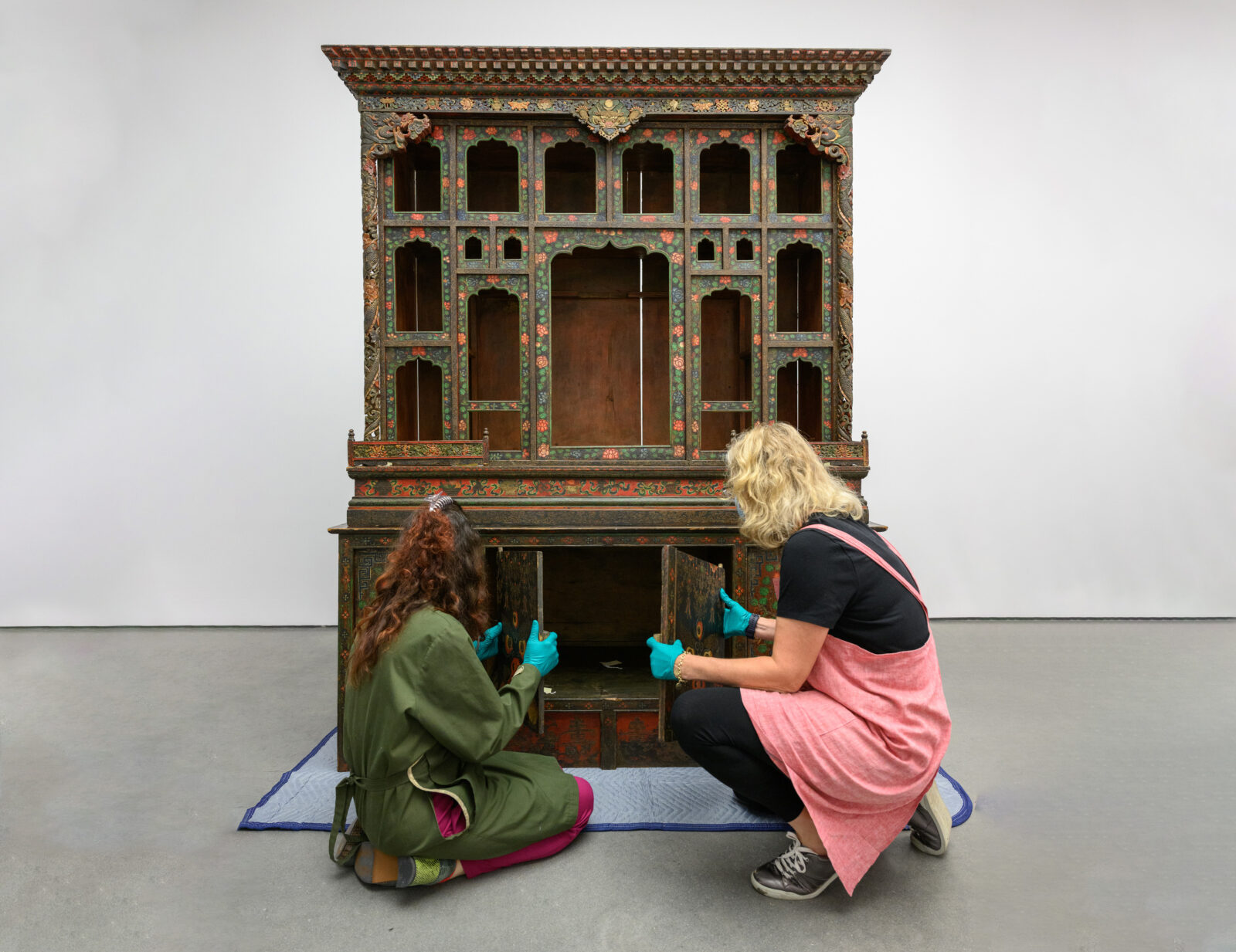 Two people with blue gloves crouching on down at the base of an ornate cabinet, putting it together.