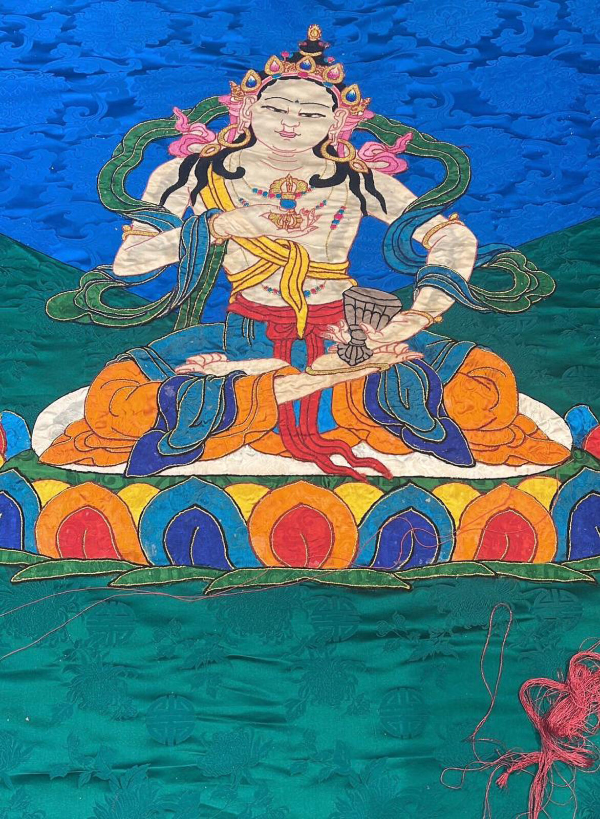 Embroidered fabric artwork of a cross-legged figure in blue, orange, and red clothing and a gold and pink headpiece, on a blue and green background