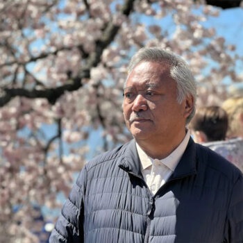 Jamyang Dorjee standing in front of a cherry blossom tree, wearing a white button down shirt and black puffer coat