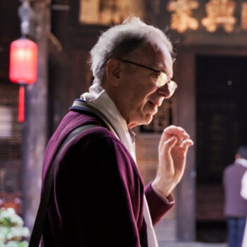 a man wearing glasses and a purple sweater raising his left hand