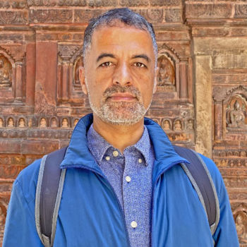 An adult man with gray hair, mustache, and beard wearing a blue jacket and button down shirt and a backpack