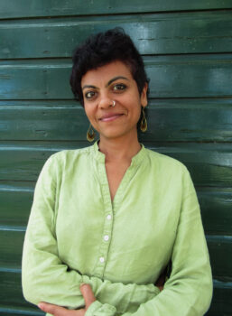 The image displays a portrait of a person with short, dark, curly hair, a nose ring, and gold earrings. They are wearing a light green, collared, button-up shirt and are standing in front of a dark green wooden wall with horizontal slats.