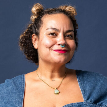 A smiling adult with blonde and brown hair wearing a denim blouse in front of a blue background