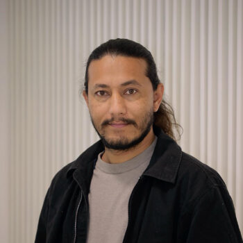Adult man with long black hair tied back, a black mustache and beard, standing in front of a gray wall