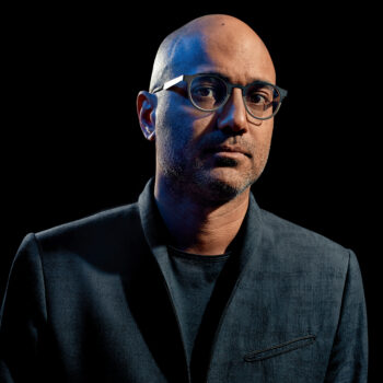 A man wearing glasses and a gray blazer stands in front of a black background