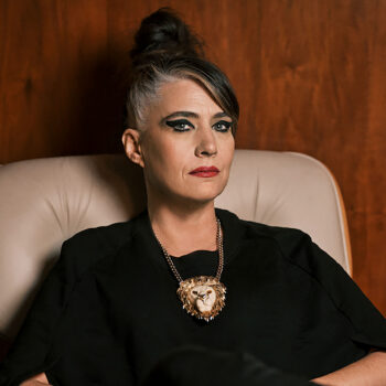 A woman wearing a lion's head necklace, black eyeliner, and red lipstick, sitting on a gray leather seat.