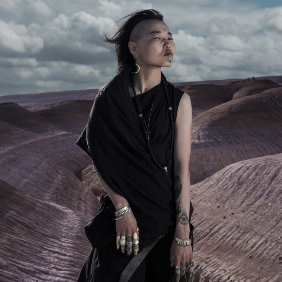 An adult woman standing in a desert landscape, wearing an all-black outfit with silver jewelry on both hands