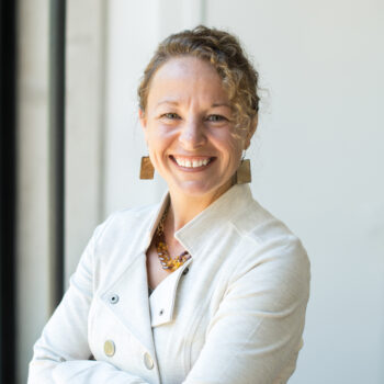 a smiling adult woman with curly blonde hair wearing a white jacket