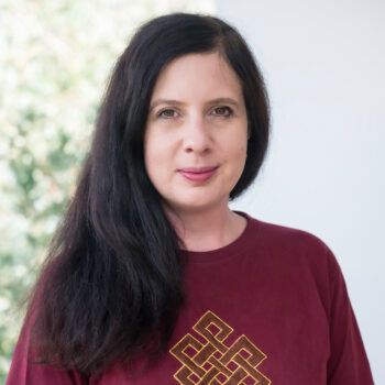 Head-and-shoulders portrait of a woman with long dark hair, wearing a maroon top with a gold geometric embroidery, against a softly blurred green-and-white background.