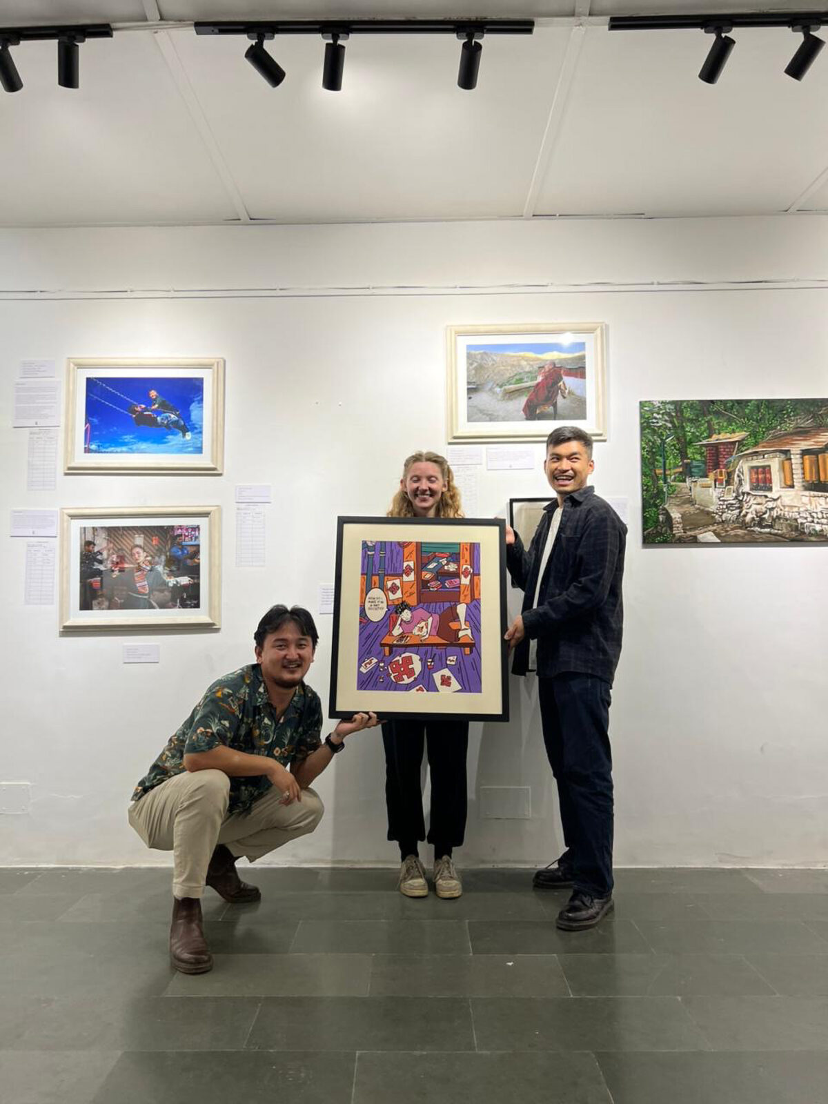 three adults hold a framed artwork in front of a white wall with additional artworks displayed