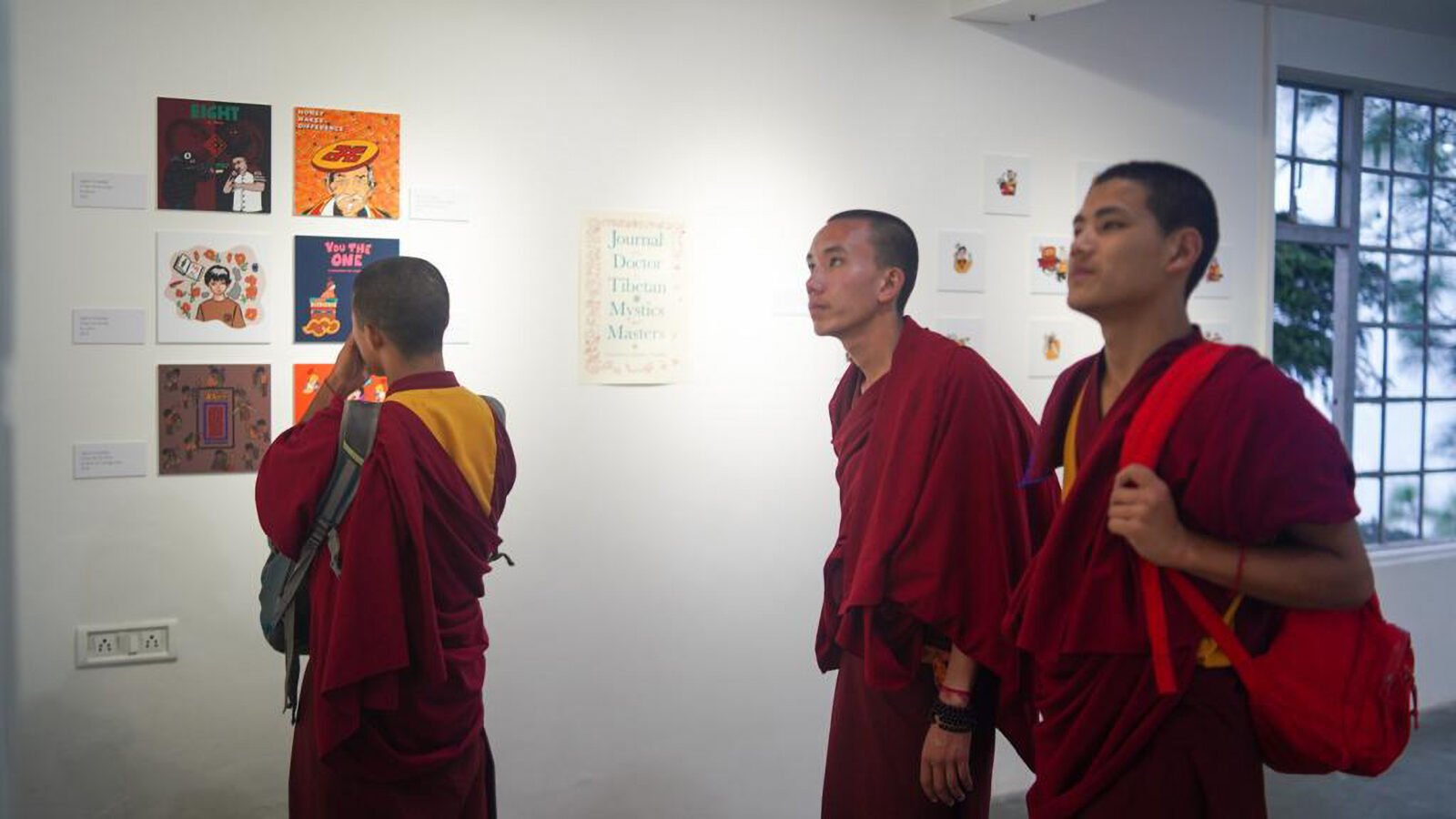 three adults in red and yellow robes look at artwork displayed on white gallery walls