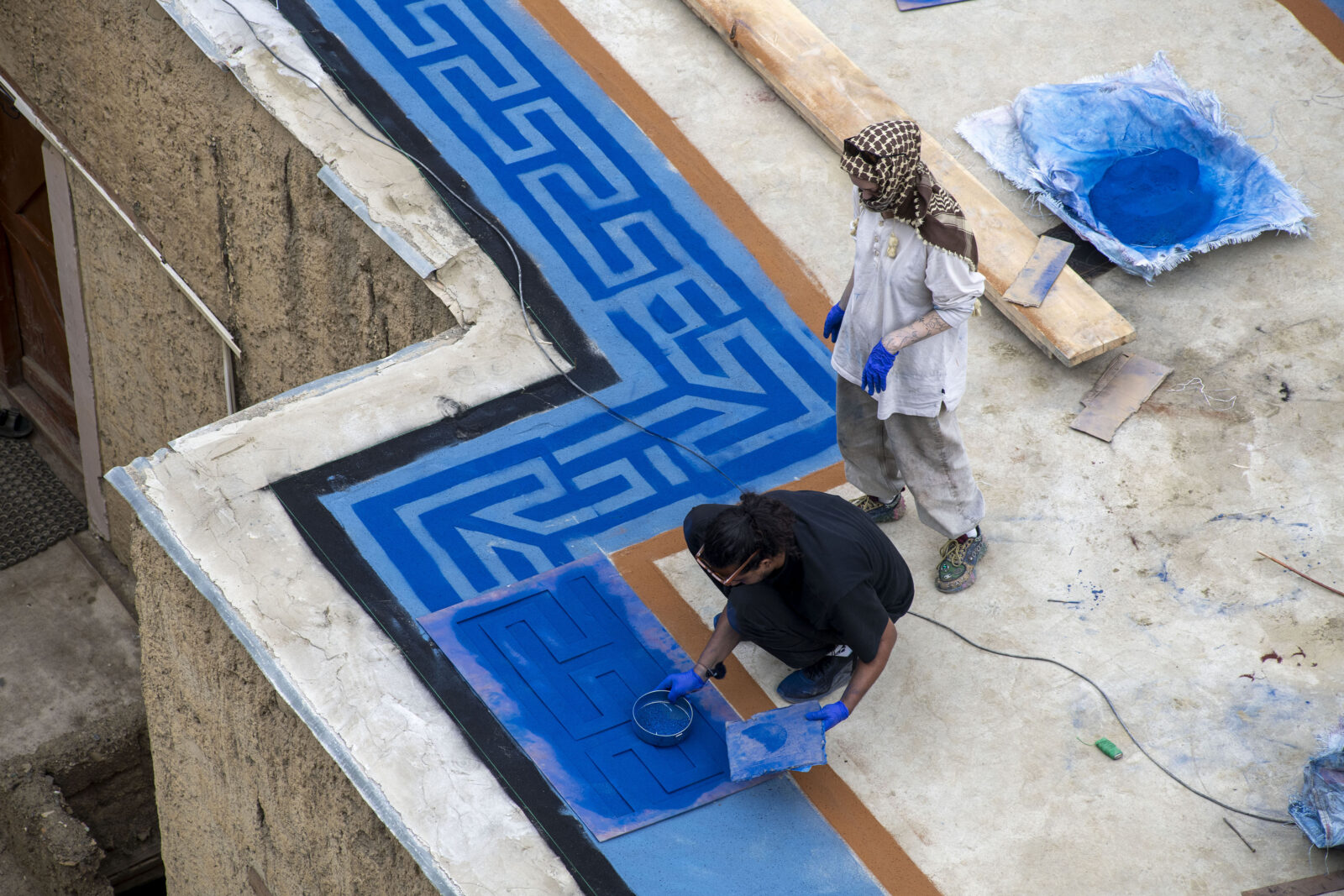 Two figures on a rooftop, with one crouching and spreading blue pigment across the roof to create a pattern while the other stands behind them and looks down at the design