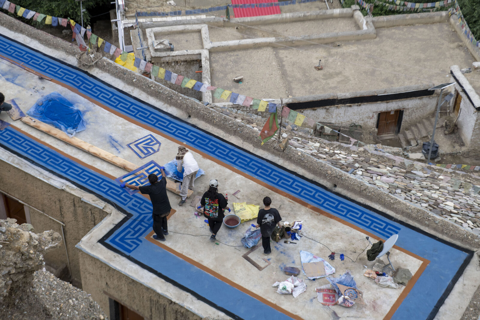 An overhead view of four figures standing on a rooftop with a blue border in progress. One figure holds up a stencil, while pigments, stencils, and other art materials are scattered across the roof