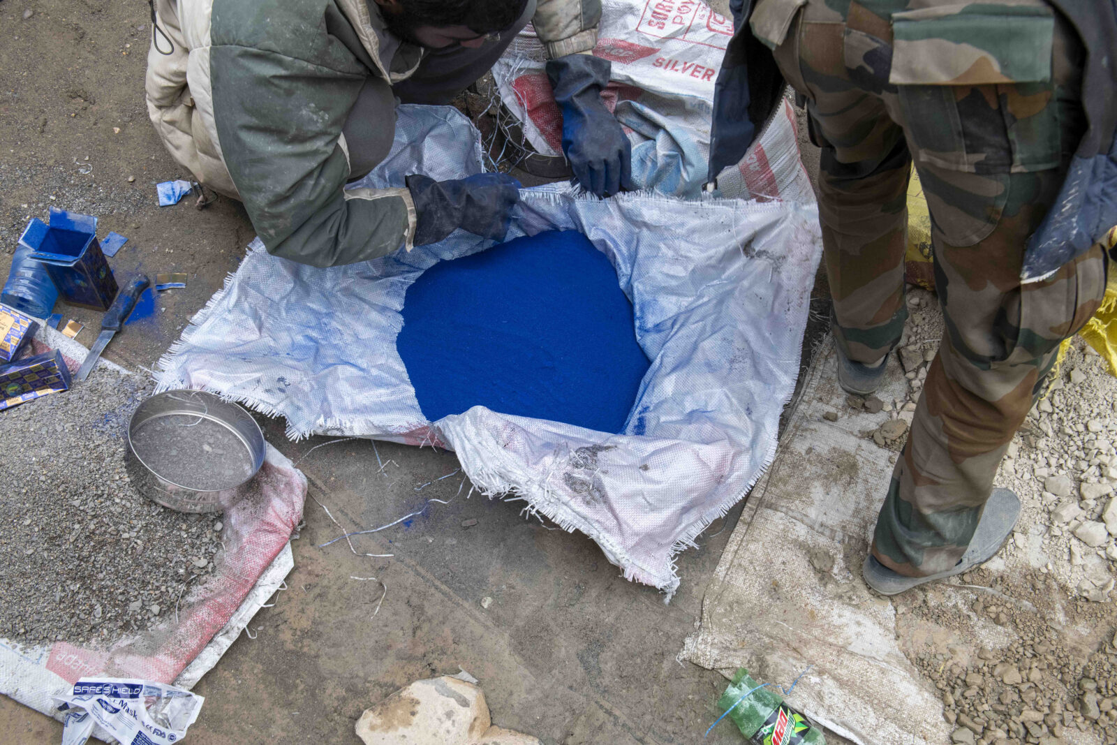 A figure opens a sack of blue pigment on the ground while another figure stands to the right