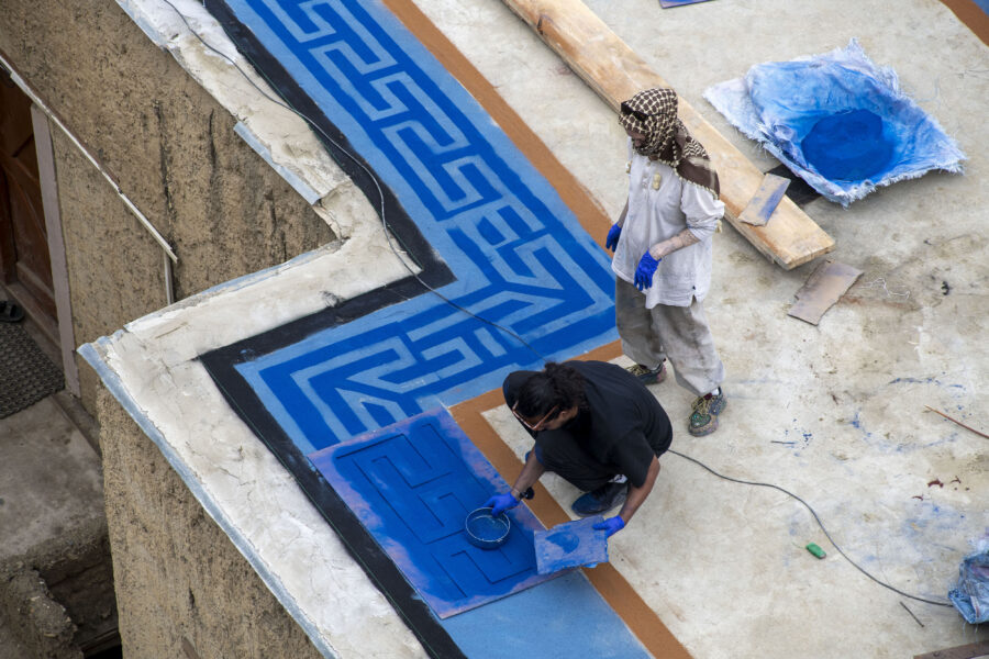 Two figures on a rooftop, with one crouching and spreading blue pigment across the roof to create a pattern while the other stands behind them and looks down at the design