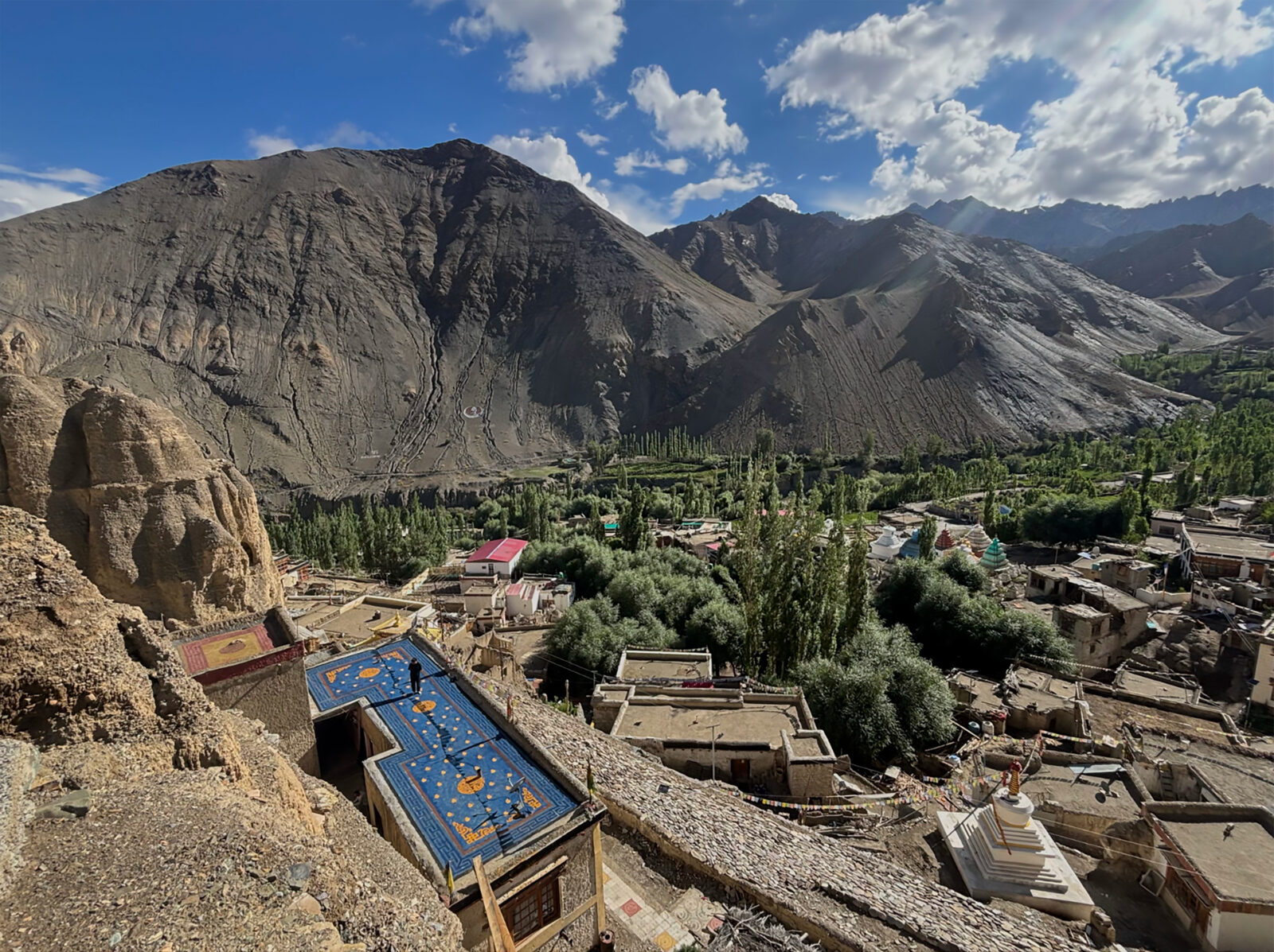 An aerial view of a mountainous area in Ladakh, India, with a painted blue and red rooftop in the foreground, trees near the base of the mountains, and mountains in the background