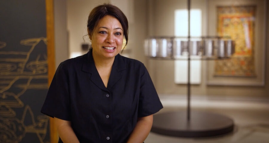 Half-length portrait of woman in blue shirt with artworks in background.