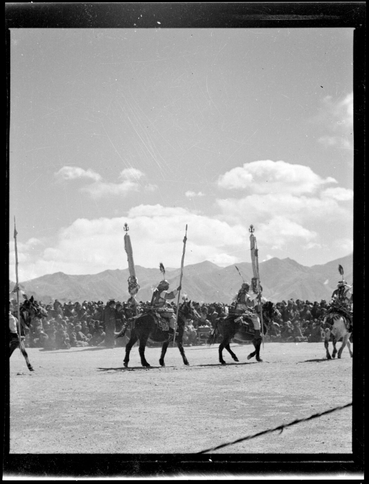 black and white photograph of four people riding horses, each holding a tall stick, in front of a large crowd