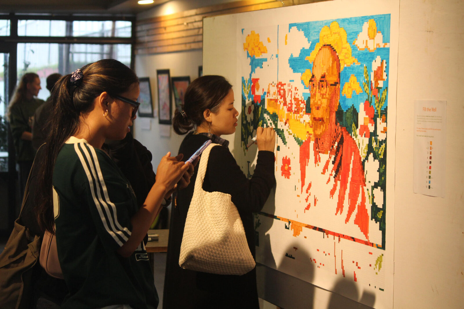 Two women stand in front of a wall-mounted artwork; one colors in a square with a marker while the other looks at her phone.