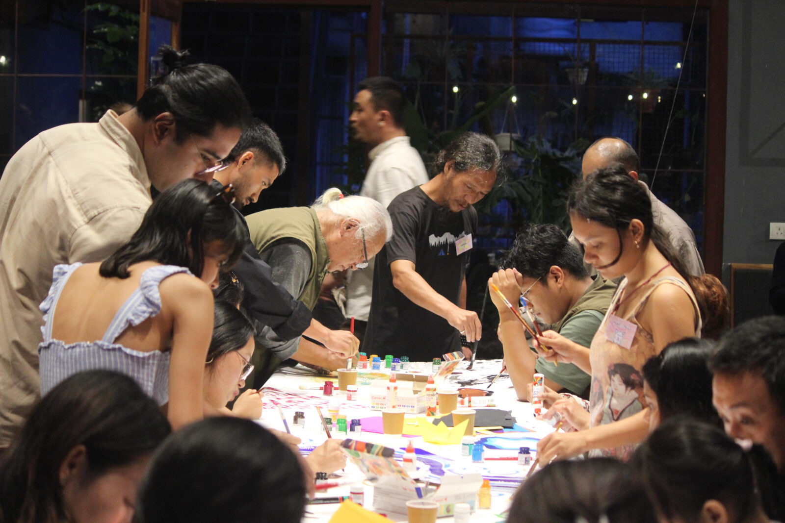 a group of people work around a table with paper, paint, paintbrushes, and glue, painting and gluing paper scraps together