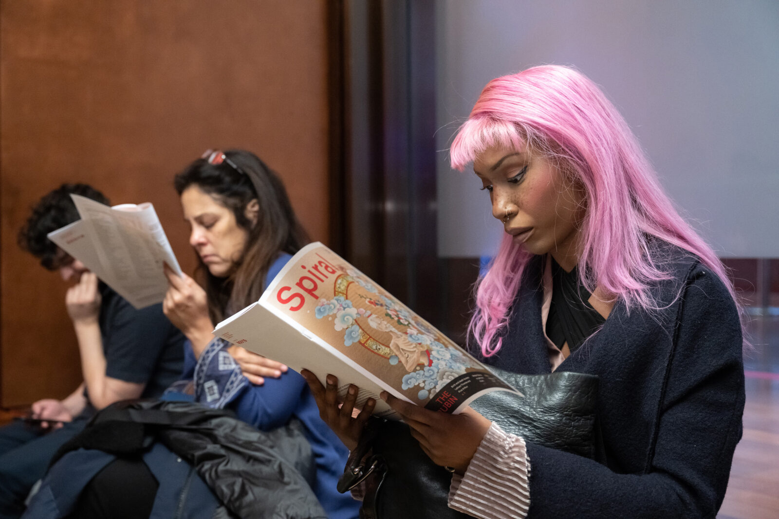 an adult with pink hair and a black coat reads a magazine titled "SPIRAL"
