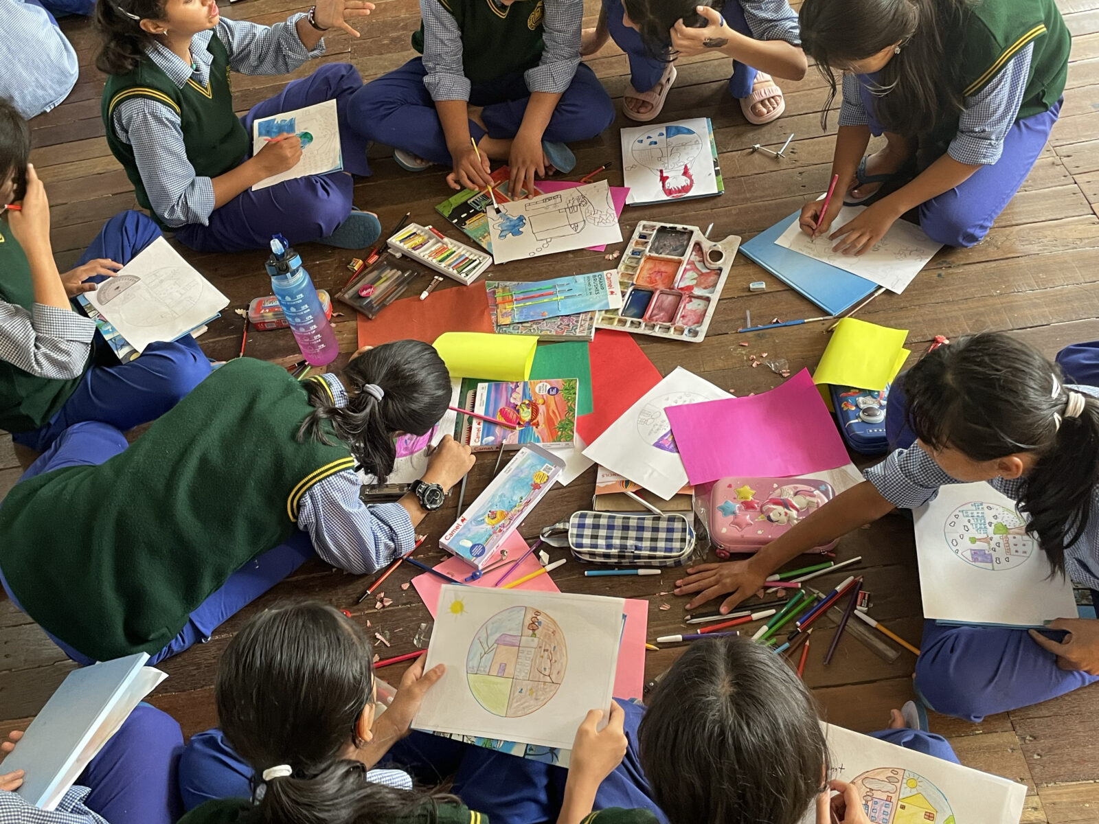 a group of students sit on the floor, using colored pencils and watercolor to draw on white and colored paper