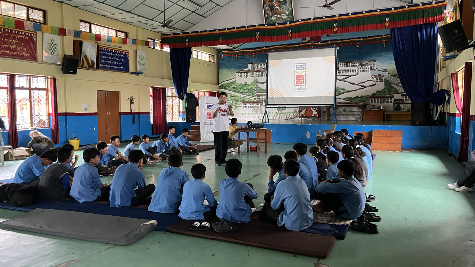A group of students sit on the floor facing a screen displaying a logo and the word "Khadhok," while a man stands between the screen and the students and speaks to them