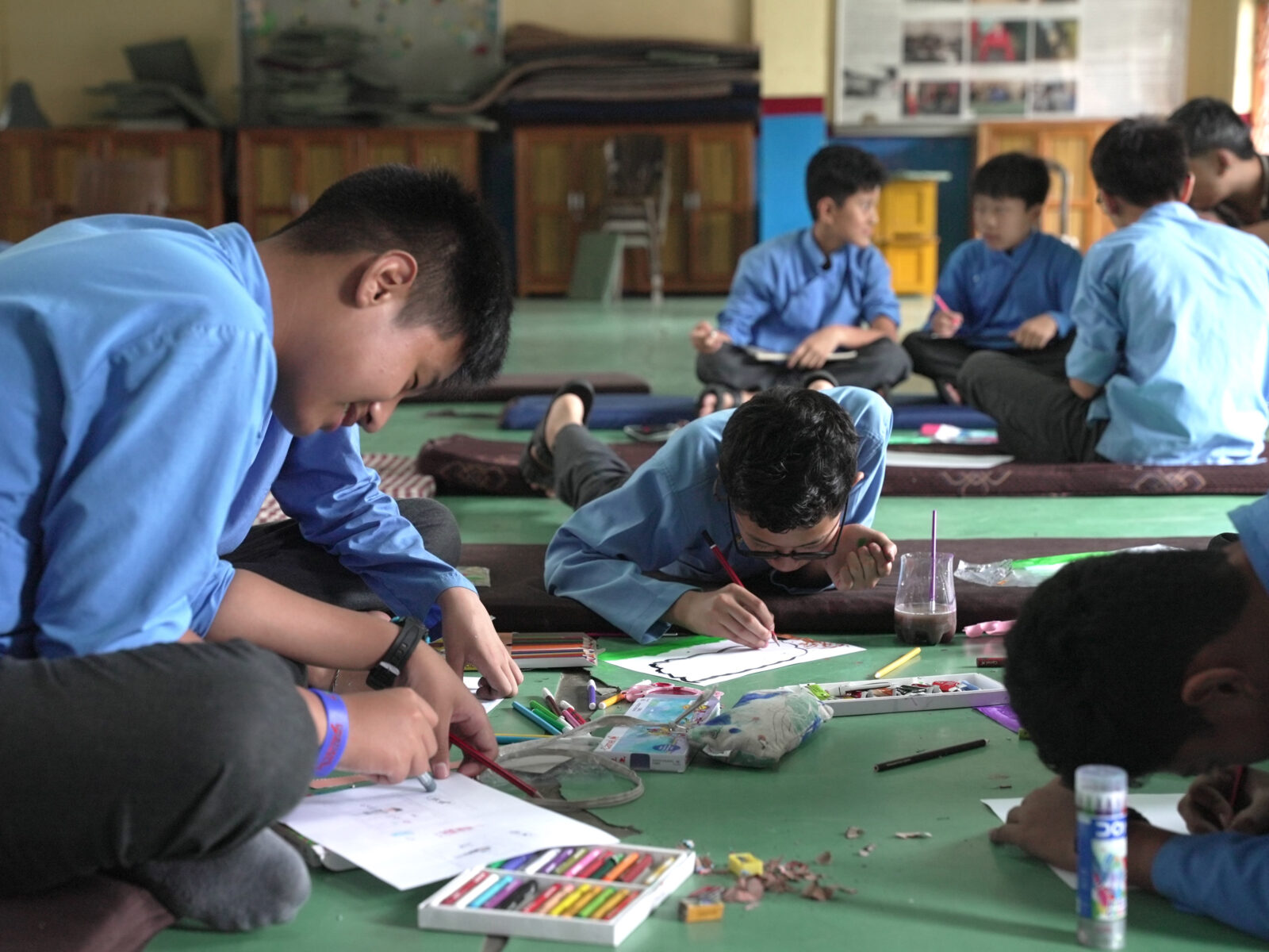 students sit on brown mats on a green floor, coloring on white paper