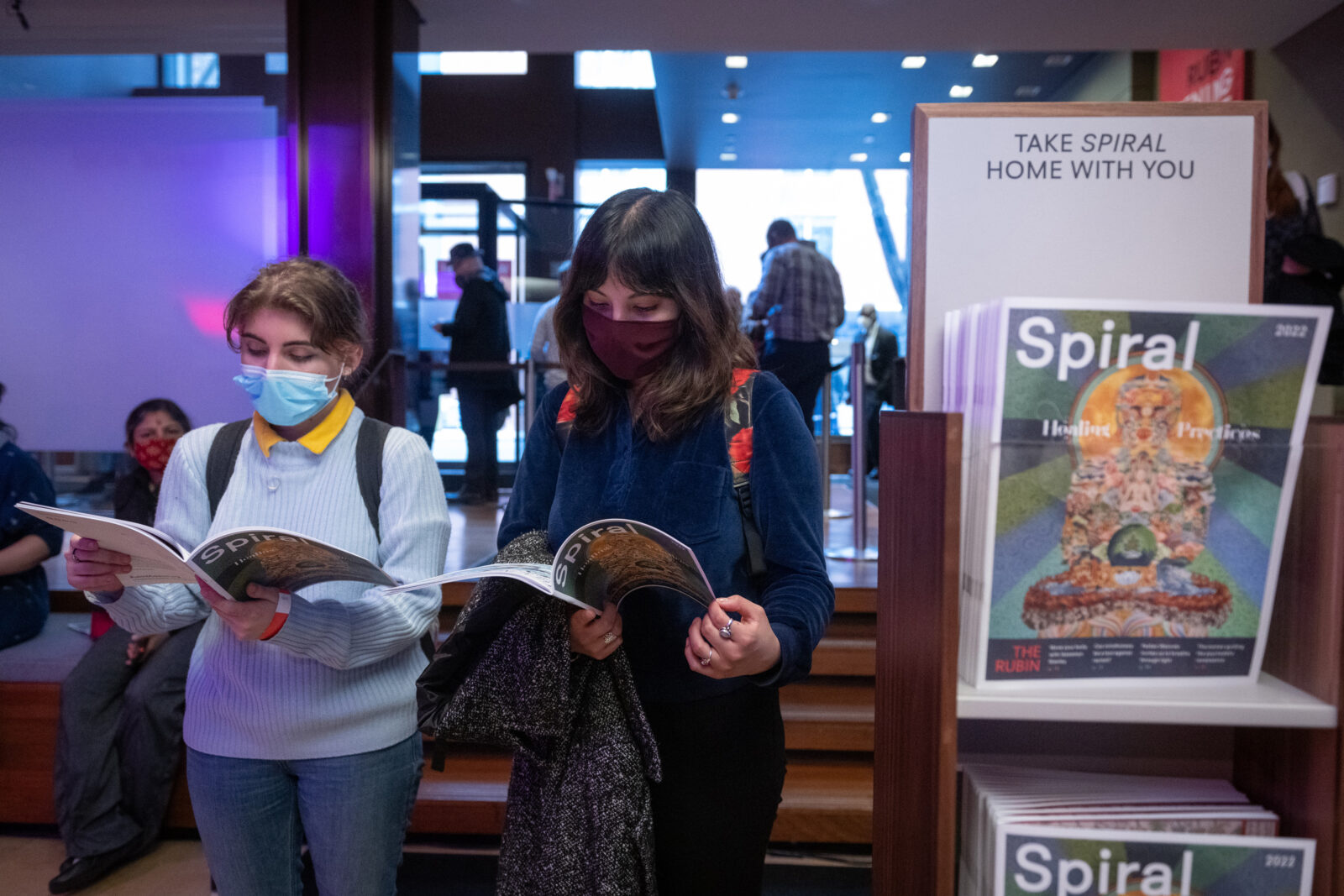 Two adults hold and read a magazine titled "Spiral." To their right, a magazine stand displays copies of magazines with the title "Spiral Healing Practices" with a sign reading "TAKE SPIRAL HOME WITH YOU."