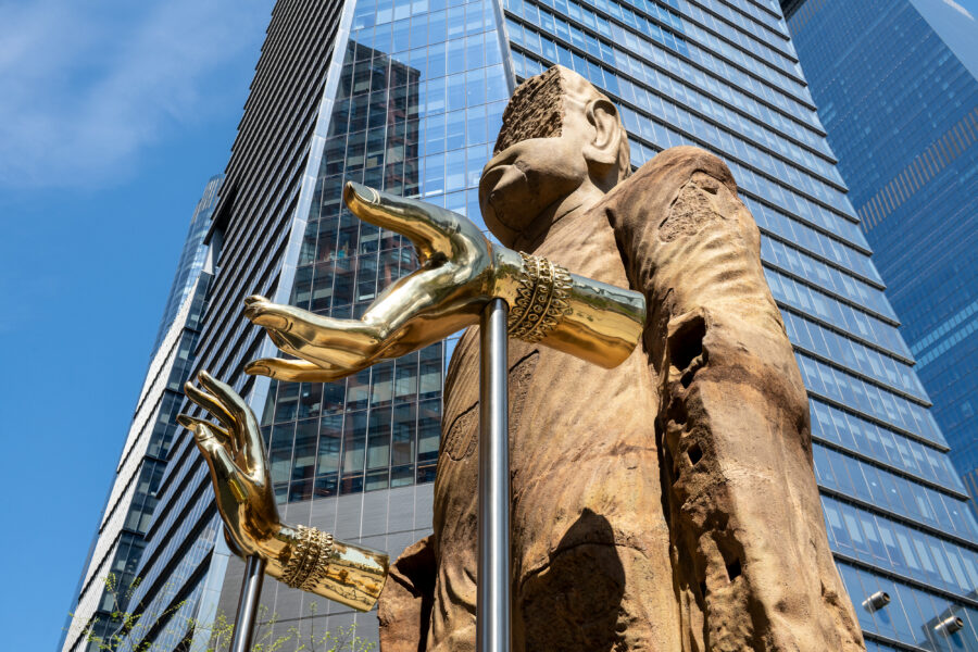 Low angle photo of a Bamiyan Buddha sculpture with gold hands in front of a tall skyscraper