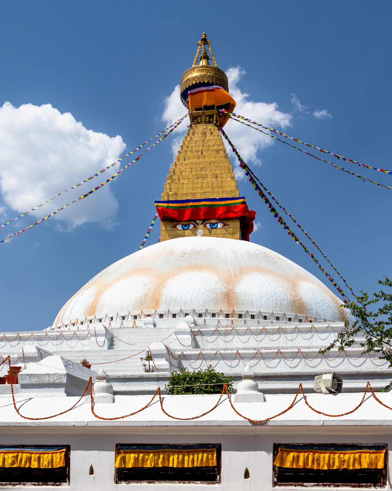 Building with multi-level rectangular base, a large white dome, and a golden spire at the center. A pair of half-lidded, blue eyes painted at the base of the spire. Strings of colorful prayer flags stream down from an umbrella-shaped canopy at the top.