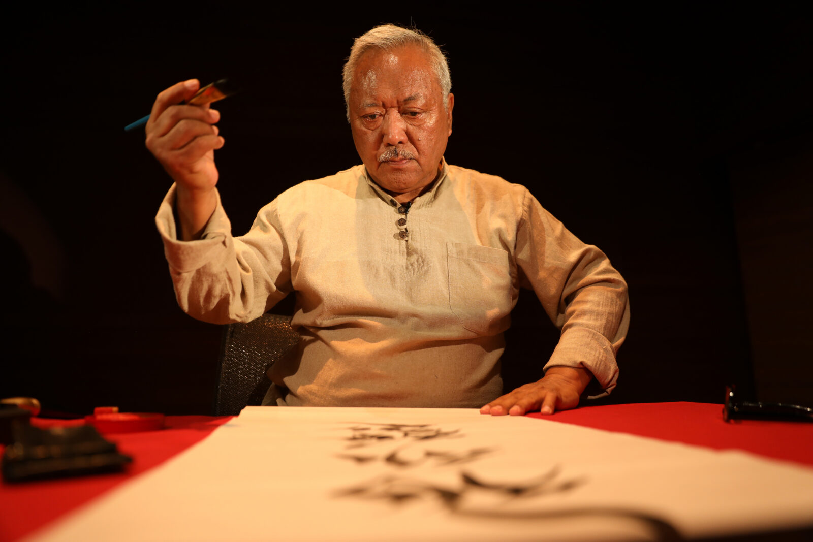 an adult man with gray hair holds a paintbrush on one hand while looking down a scroll on a red table