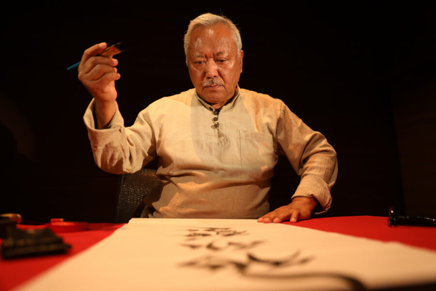 an adult man with gray hair holds a paintbrush on one hand while looking down a scroll on a red table