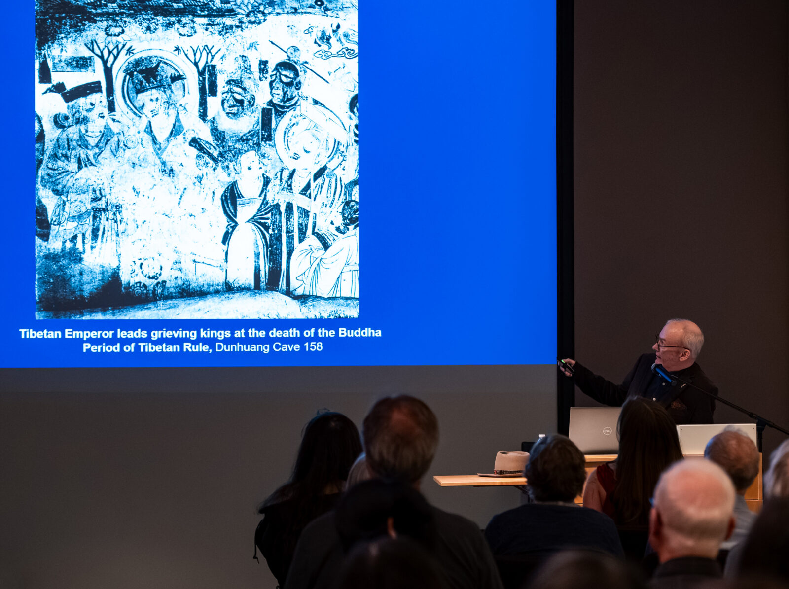 Karl Debreczney stands to the side of a screen displaying an image of an artwork with the caption, "Tibetan Emperor leads grieving kings at the death of the Buddha, Period of Tibetan Rule, Dunhuang Cave 158"
