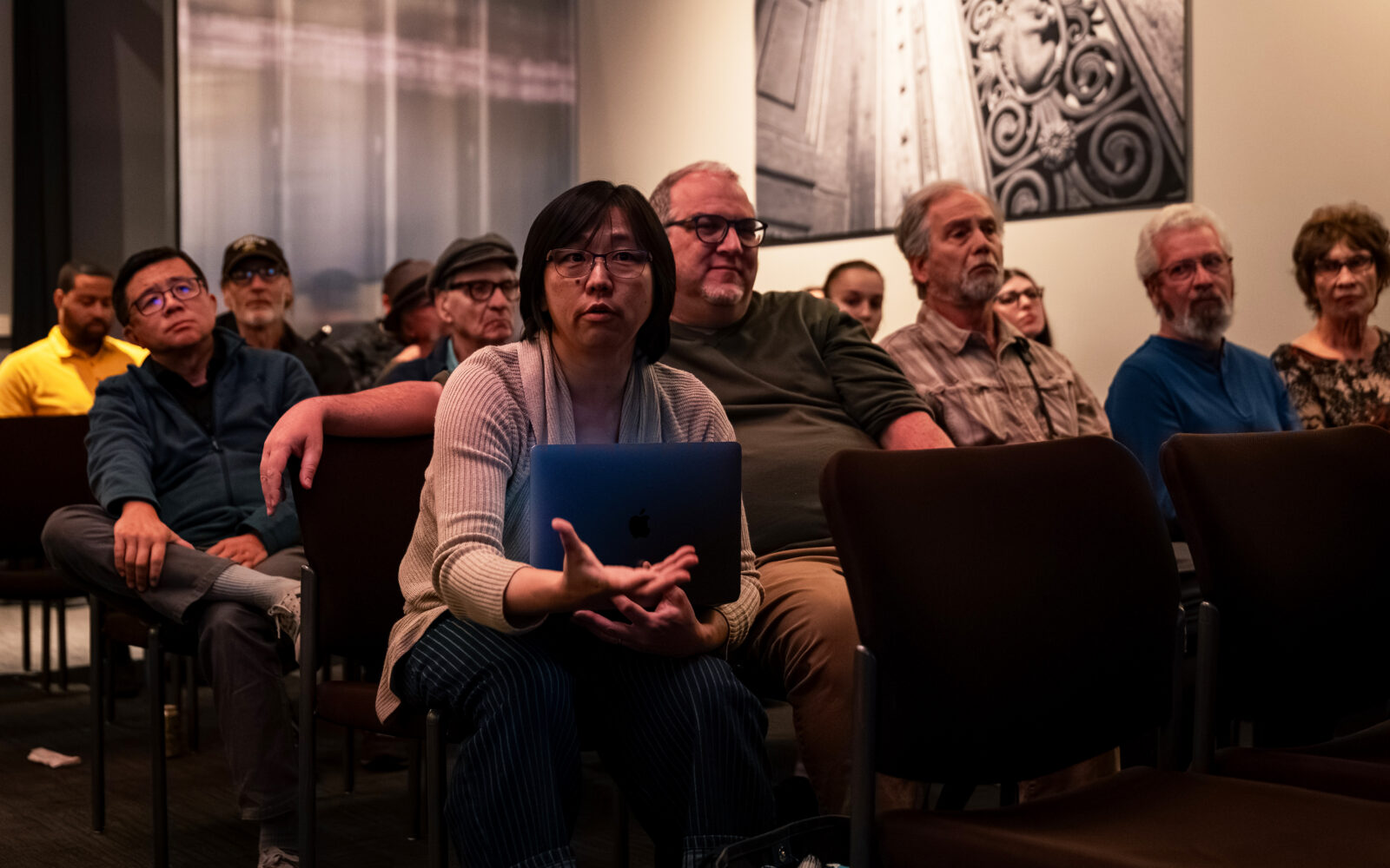Audience members seated with an adult woman at the front leaning forward and talking while holding a laptop