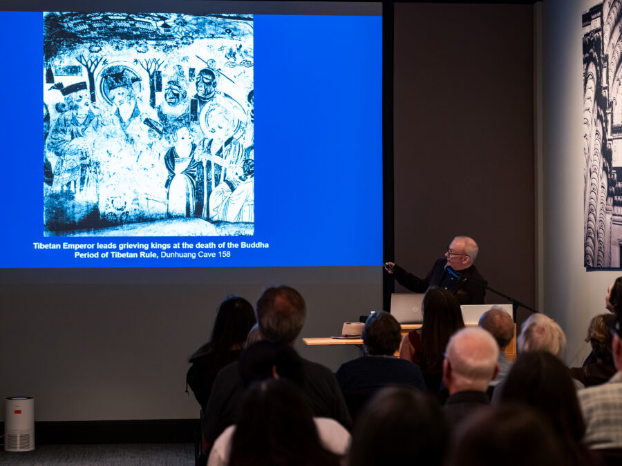 Karl Debreczney stands to the side of a screen displaying an image of an artwork with the caption, "Tibetan Emperor leads grieving kings at the death of the Buddha, Period of Tibetan Rule, Dunhuang Cave 158"