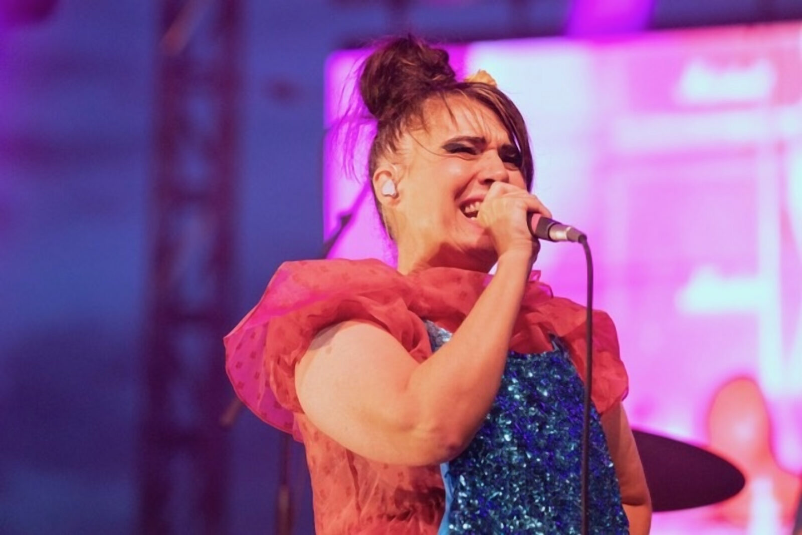 An adult wearing a pink and blue dress singing with eyes closed while holding a mic close to their mouth